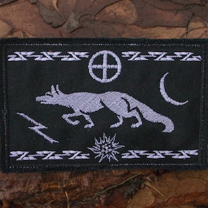 May include: A rectangular black embroidered patch featuring a light gray running fox at the center. The design includes a sun cross, a crescent moon, a lightning bolt, and a star, framed by horizontal borders with repeating geometric patterns.