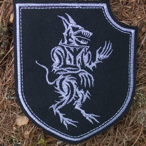 May include: A black embroidered patch in the shape of a shield with a white stylised wolf design.
