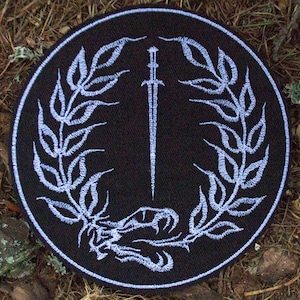May include: A round black patch with a white border. The patch features a white sword design in the center, surrounded by a laurel wreath. A snake is at the base of the sword. The patch is set on a bed of brown leaves and twigs.