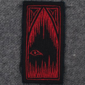 Könnte beinhalten: Schwarzer und roter gestickter Patch mit einem stilisierten Auge und Flammen. Der Patch ist rechteckig und hat einen ausgefransten Rand.