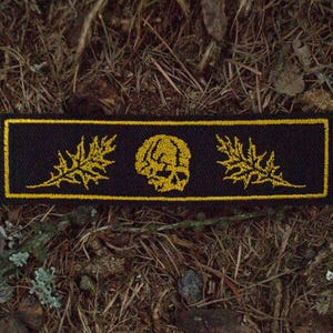 May include: Black rectangular patch with a gold border, featuring a gold skull in the center and gold leaf designs on each side. The patch is set against a backdrop of brown and green foliage.