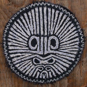 Może przedstawiać: Okrągła, czarno-biała, haftowana naszywka z wystylizowanym wzorem twarzy. Twarz ma duże oczy, nos i usta, z promienistymi liniami rozchodzącymi się na zewnątrz. Naszywka znajduje się na drewnianej powierzchni.