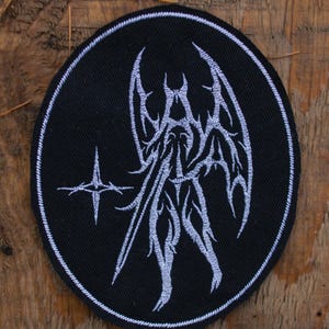 May include: Black circular patch with a white embroidered design featuring a stylized figure with wings and a cross-like symbol. The patch is set against a wooden background.
