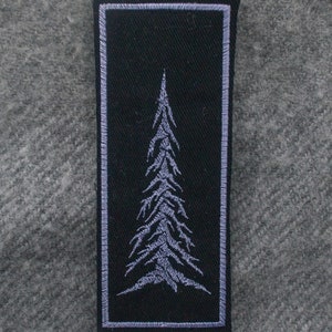May include: A rectangular black patch with a detailed embroidered tree design in light gray. The patch has a light gray border and is set against a gray fabric background. The tree design is centered on the patch.