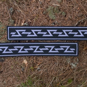 May include: Two black fabric strips with a repeating geometric pattern in a light purple colour. The pattern is a series of zigzags.