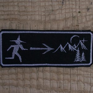 Può includere: Toppa rettangolare nera con bordo bianco. La toppa presenta una figura che corre con un cappello da strega, una freccia e un paesaggio montano con luna crescente e pini. Su una superficie di legno.