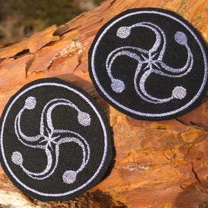 May include: Two circular black patches with a white embroidered design. Each patch features a four-armed spiral within a white circular border. The patches are shown on a textured, brown surface.