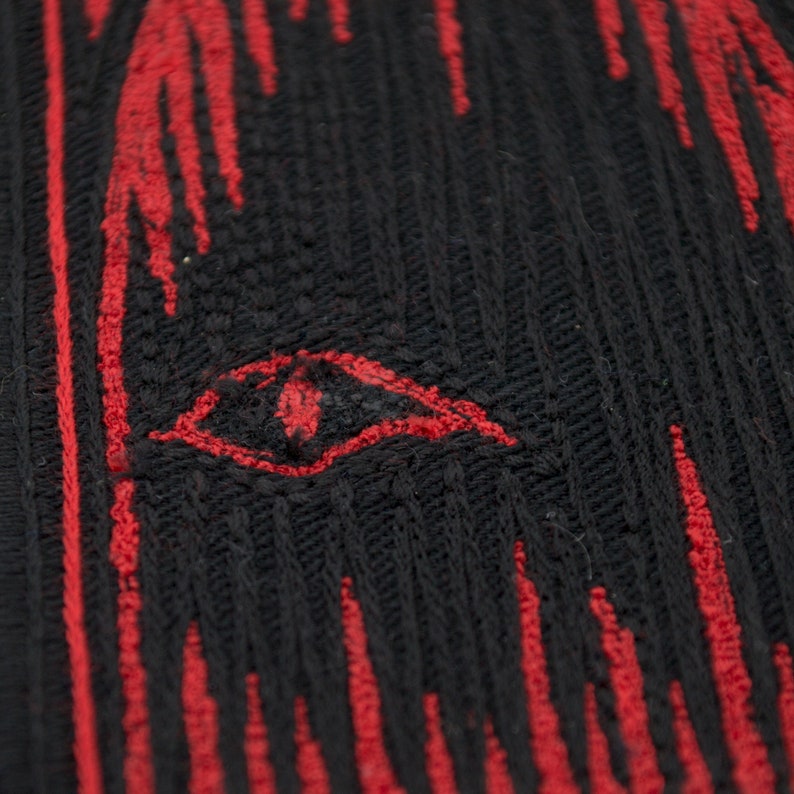 Pode incluir: Close-up de um tecido preto com listras verticais vermelhas. Uma forma de olho vermelha &eacute; bordada no centro do tecido.