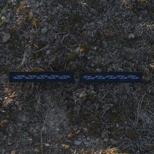 May include: Two black rectangular patches with a geometric design in white and light blue. The patches are lying on a bed of soil and small plants. The design is a repeating pattern of zigzags and lines.