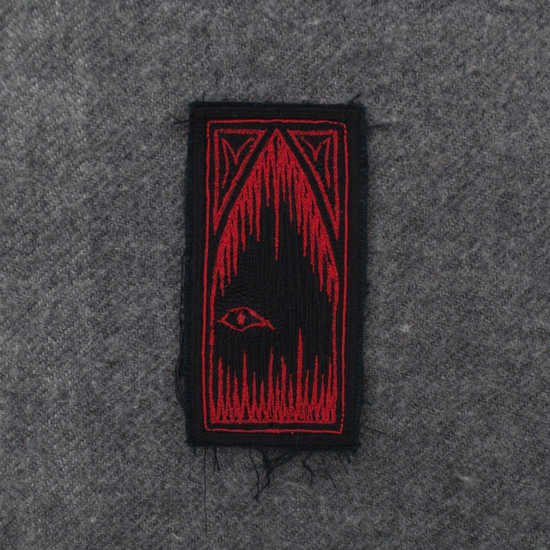 Pode incluir: Etiqueta bordada preta e vermelha com um olho estilizado e chamas. A etiqueta &eacute; retangular e tem uma borda desfiada.