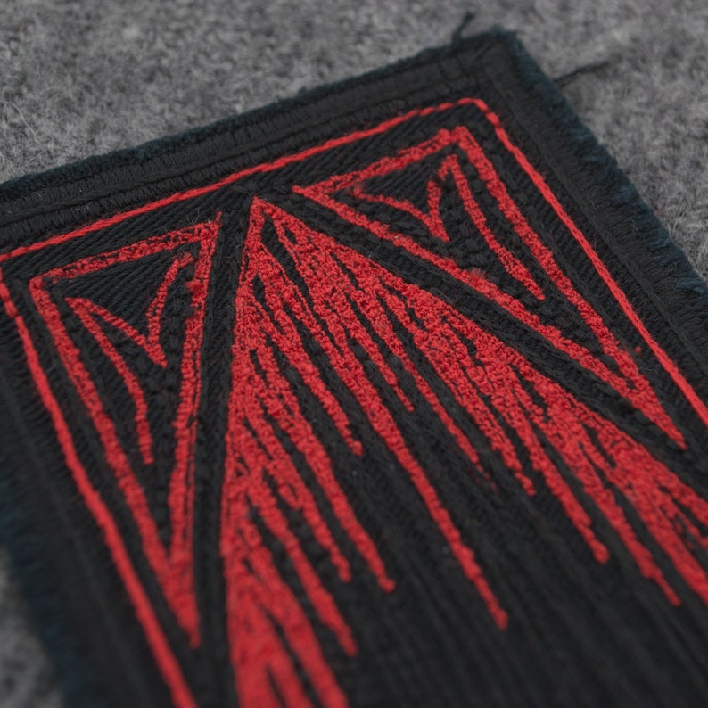 Pode incluir: Um close-up de um remendo de tecido preto com um design bordado vermelho. O design apresenta uma s&eacute;rie de linhas verticais que criam um padr&atilde;o geom&eacute;trico.