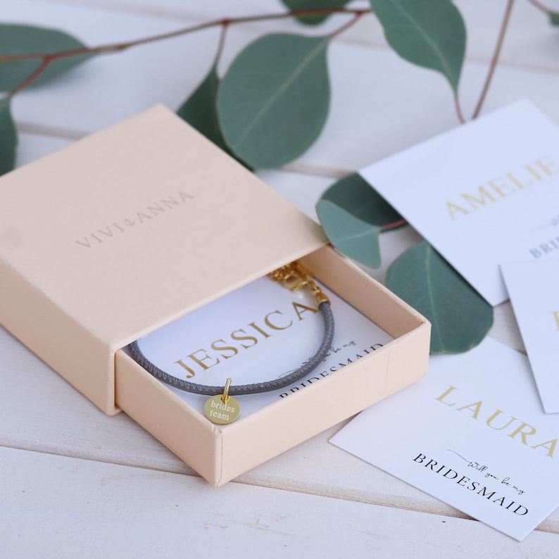 May include: A gray bracelet with a gold charm that says "brides team" in a light pink box with the text "VIVIANNA" on the top. The box is sitting on a white wooden surface. There are also white cards with the text "AMELIE BRIDESMAID", "LAURA will you be my BRIDESMAID" and "JESSICA BRIDESMAID" on the surface.