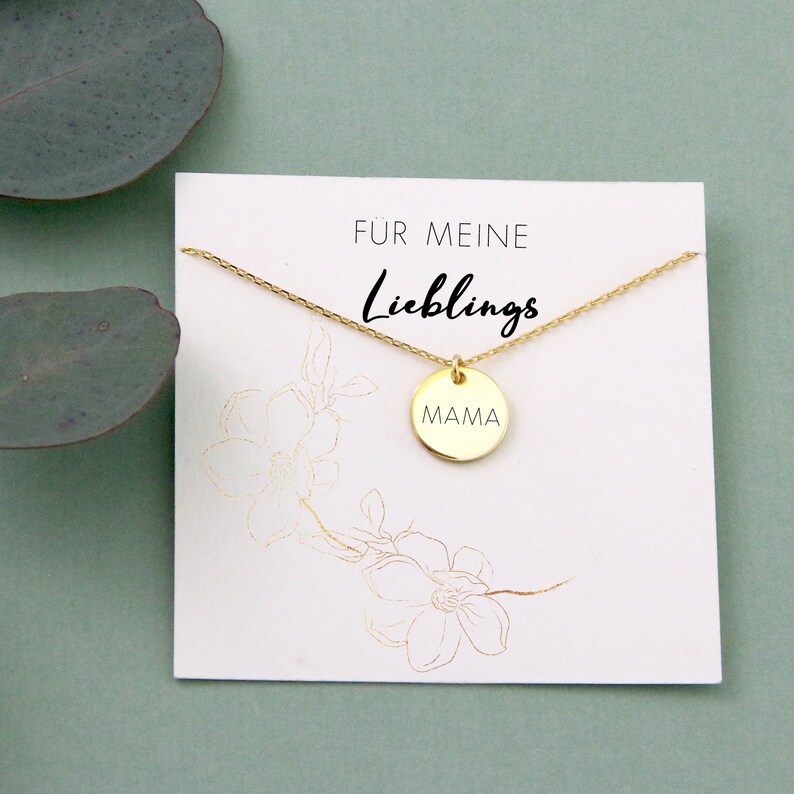 May include: Gold necklace with a round pendant that says "MAMA" on it. The necklace is on a white card with the text "F&uuml;r meine Lieblings" on it.