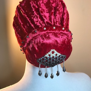 May include: A red velvet bun with a beaded embellishment. The bun is decorated with small white pearls and black beads. The embellishment features a row of black beads with a row of white pearls above it. The embellishment has a dangling row of black beads.