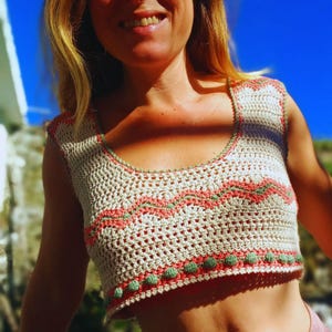 Puede incluir: Un top corto de crochet con base blanca y detalles en rosa y verde. El top tiene un borde festoneado y una trama suelta y abierta.