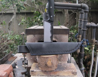 May include: A black leather sheath with a black knife with a silver blade is resting on a brick and mortar structure. The knife is resting on a wooden block. The knife is partially inserted into the sheath. The background is a wooden fence and a green bush.