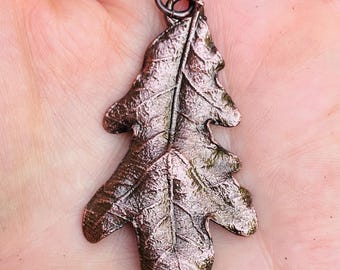 May include: A bronze-colored leaf pendant with intricate veining details. The pendant is shaped like an oak leaf and has a small loop at the top for a chain. The metal has a textured surface and a dark, antique finish.