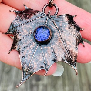 May include: A copper maple leaf pendant with a purple glass stone in the center. The pendant is hanging from a copper chain.