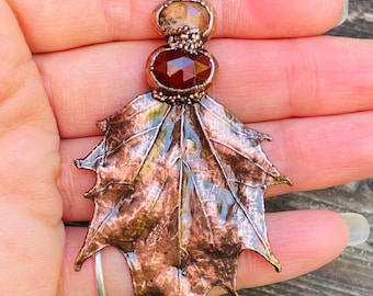 May include: Copper pendant featuring a detailed leaf design with a central oval-shaped, deep red gemstone and a smaller, light-colored stone above. The pendant is held by a hand, showcasing its intricate metalwork.