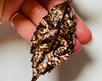 May include: A copper-colored leaf pendant with a textured surface. The pendant is attached to a silver chain.