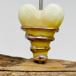 May include: A heart-shaped glass bead featuring a pale yellow top and a vibrant yellow base. The bead is wrapped in a metallic, iridescent copper-colored glass coil that spirals around the lower section. A silver metal pin extends from the top.
