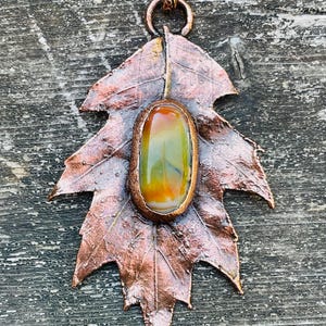 May include: A copper-colored leaf pendant with a central, oval-shaped stone. The stone features shades of green, yellow, and orange. The pendant has a detailed leaf design and a decorative loop for hanging. The pendant is approximately 3 inches long.