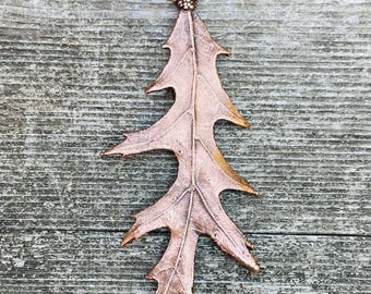 May include: A copper-colored leaf pendant with intricate veining and a textured surface. The pendant has a loop at the top for hanging and is set against a weathered wooden background. The leaf design is detailed and realistic.
