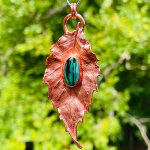 May include: A copper leaf pendant with a green and blue oval stone set in the center. The pendant hangs from a silver chain against a blurred green background. The leaf has detailed veining.