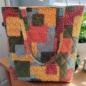 May include: A quilted tote bag with a patchwork design. The bag features squares of red, yellow, blue, and green fabric with floral and polka dot patterns. The bag has a quilted texture and sturdy handles.