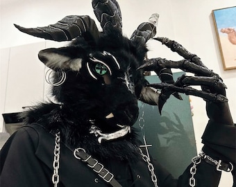 Op de afbeelding: Een zwart, bont masker met hoorns en een zilveren ketting. Het masker heeft groene ogen en een zilveren mond. De persoon die het masker draagt, draagt ook een zwart jack met zilveren kettingen en een zwarte riem.