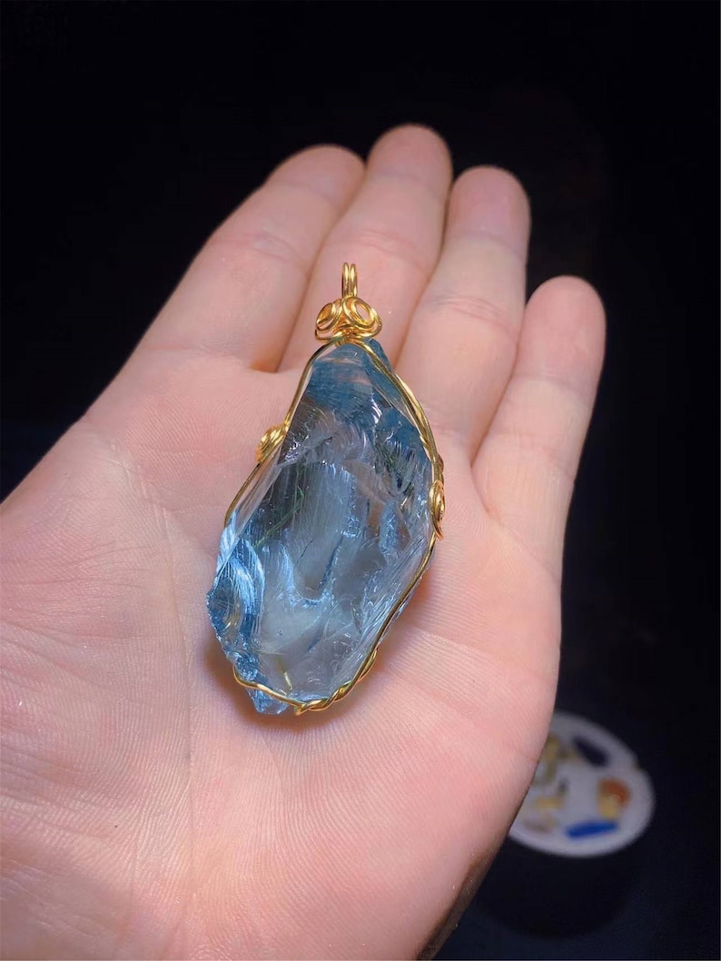 Op de afbeelding: Een lichtblauwe kristallen hanger gewikkeld in gouden draad. De kristal is gefacetteerd en heeft een natuurlijke, ruwe vorm. De hanger hangt aan een gouden ketting.