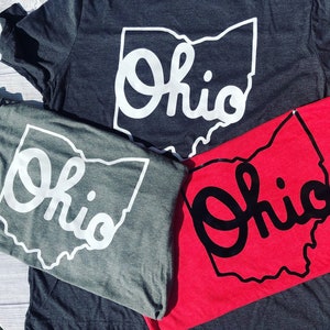 May include: Two t-shirts with the word "Ohio" in a stylized font inside the outline of the state of Ohio. One shirt is gray with white lettering and the other is red with black lettering.