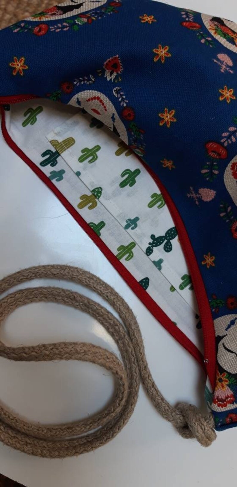 May include: A blue fabric bag with a white lining and a red zipper. The bag has a floral pattern with cacti and a rope strap.
