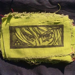 May include: A green fabric patch with a black and white image of an anime character's eye. The image is framed in a black rectangle.
