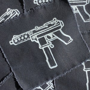 May include: A collection of black fabric patches with a white outline of a machine gun. The patches have a square shape with frayed edges. The image is a close-up shot of the patches.