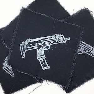 May include: Several black fabric patches with a white outline of a submachine gun design. The patches have a square shape with frayed edges. The image is taken from a slightly elevated angle, showing the patches overlapping.
