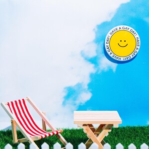 May include: A miniature white picket fence in front of a red and white striped deckchair and a small wooden table on a green grassy surface. A yellow smiley face button with the text "Have a Gay Day!" is in the blue sky above.