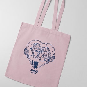 May include: A pink tote bag featuring a blue illustration of two people kissing within a heart adorned with stars. The text "ANNIE goes blue" is printed beneath the heart.