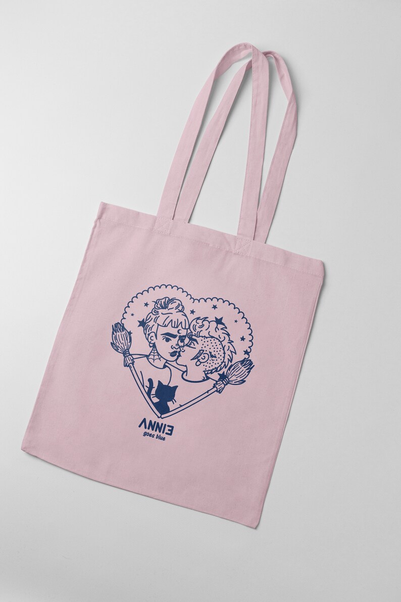 May include: A pink tote bag featuring a blue illustration of two people kissing within a heart adorned with stars. The text "ANNIE goes blue" is printed beneath the heart.