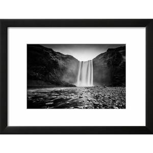 May include: A black and white photograph of a waterfall cascading down a rocky cliff face. The water is flowing over the edge of the cliff and creating a mist in the air. There are people standing at the bottom of the waterfall.
