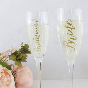 May include: Two champagne flutes with gold lettering. One flute says "Bride" and the other says "Bridesmaid".