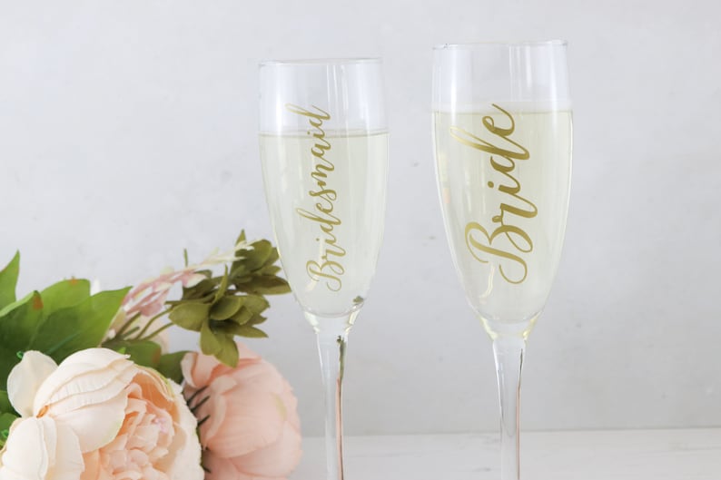 May include: Two champagne flutes with gold lettering. One flute says "Bride" and the other says "Bridesmaid".