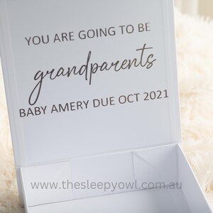 May include: A white gift box with the message "YOU ARE GOING TO BE grandparents" and "BABY AMERY DUE OCT 2021". The box is open, revealing the inside. The background is a soft, fluffy, white material.