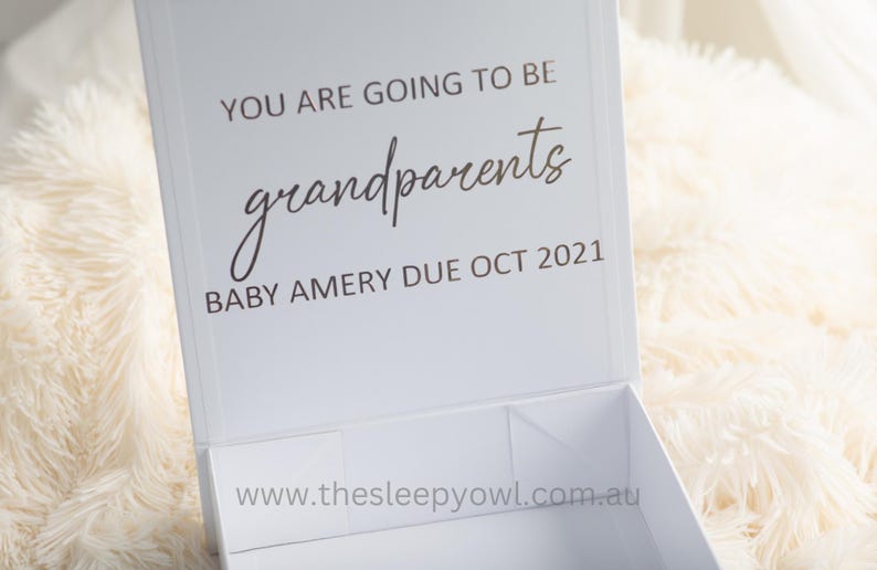 May include: A white gift box with the message "YOU ARE GOING TO BE grandparents" and "BABY AMERY DUE OCT 2021". The box is open, revealing the inside. The background is a soft, fluffy, white material.