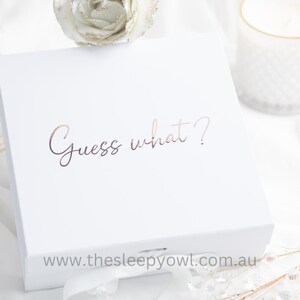 May include: A white gift box with the phrase "Guess what?" in rose gold script. The box is on a white surface with decorative elements, including a candle and floral accents. The website address www.thesleepyowl.com.au is at the bottom.