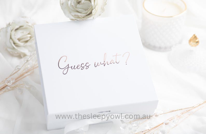 May include: A white gift box with the phrase "Guess what?" in rose gold script. The box is on a white surface with decorative elements, including a candle and floral accents. The website address www.thesleepyowl.com.au is at the bottom.