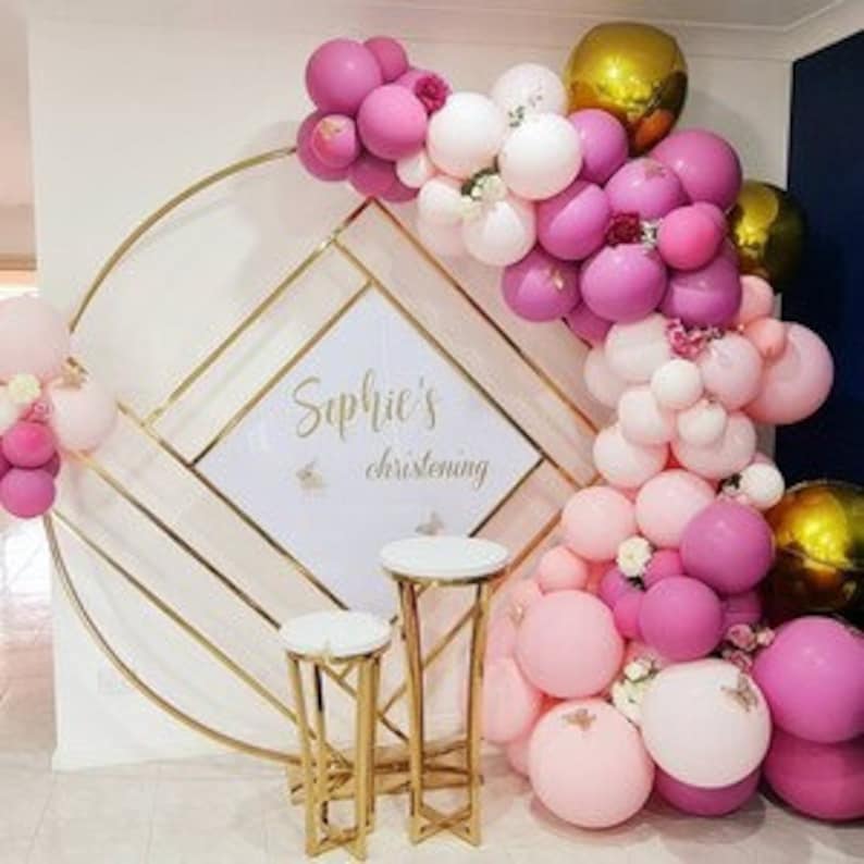 May include: A gold metal arch with a geometric design and a white backdrop with the text "Sophie's christening". The arch is decorated with pink, white, and gold balloons.