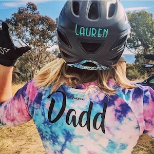May include: A person wearing a black helmet with the name "Lauren" written on it. The person is wearing a tie-dye shirt with the word "Dadd" written on the back.