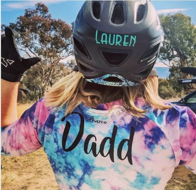 May include: A person wearing a black helmet with the name "Lauren" written on it. The person is wearing a tie-dye shirt with the word "Dadd" written on the back.