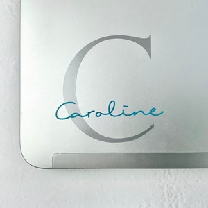May include: A silver laptop lid featuring a large, light grey capital letter C as a background decal. Overlaid on the letter is the name Caroline written in a teal, cursive script font. A small portion of a silver logo is visible in the corner.